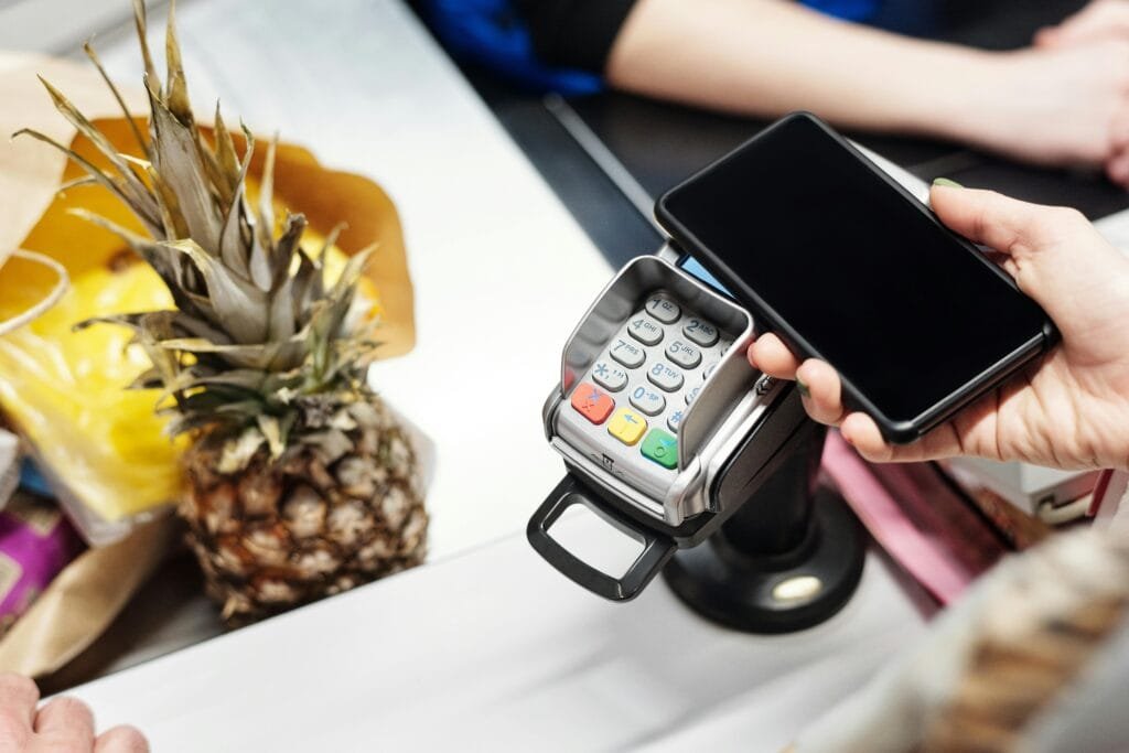 Close-up of a customer using a smartphone for contactless payment at a retail checkout with a pineapple on the counter.App financiera en viaje – control gastos viajando